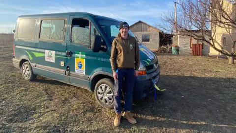 Adam Steward stood in front of a green truck which has signs on it. He is wearing a dark green jumper with blue trousers and brown boots. The vehicle is on an area of grass with a house in the background.