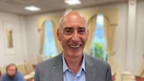 A bald man, in a light grey suit jacket, with a light blue shirt underneath, smiles at the camera. Behind him is a function room with yellow curtained windows. 