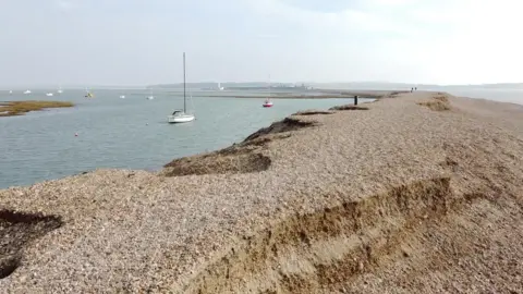 A spit of land stretches into the distance, its sides eroded by the sea. Sailboats are in the sea to the left of the land.