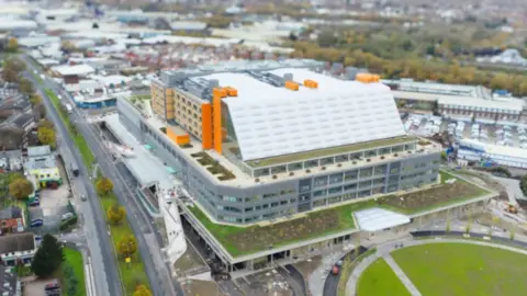 Sandwell and West Birmingham NHS Trust Aerial view of a very large rectangular building, with grey floors at the bottom, and a white roof with orange elements to the side and top.