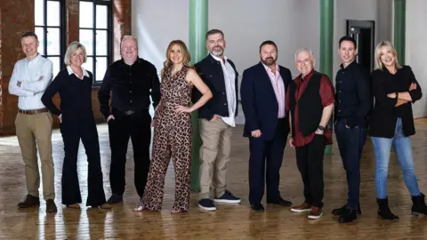 Presseye A line up photo featuring nine Radio Ulster presenters, all dressed semi-casual, pictured in a large, bright room with windows in background