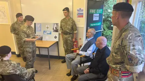 Hereford and Worcester Army Cadet Force An elderly man is sat as a group of young people in military uniform are taking part in a presentation in his honour. There is a man in a black shirt sat next to the older man, and another man in military uniform is stood in the foreground.