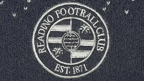 The image shows a close up of the Reading Football Club's badge on a grey woollen hat. The badge is grey and white. Instead of saying "READING FOOTBALL CLUB" the writing in the badge says "READINO FOOTRALL CLU8".
