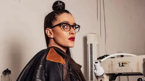 A woman with dark hair and glasses, with a bun on the top of her head, sitting in a room with manufacturing equipment and wearing a black leather jacket