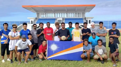 Federación de fútbol de Marshall Islands Un equipo de hombres jóvenes posa en un campo con una pelota de fútbol y la bandera de las Islas Marsahall, que es azul, con una estrella blanca y rayas diagonales blancas y naranjas. Hay una pequeña posición deportiva detrás de ellos y el mar más allá.