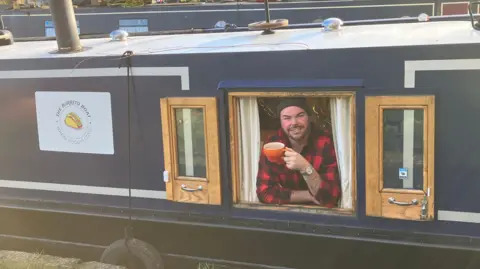 BBC/Dan Sambell A man drinking from an orange mug looks out of the window of his navy painted narrow boat. He is wearing a red and black lumberjack shirt and black hat. A painted sign for the burrito boat is to his right on the boat.