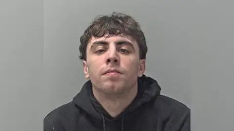 A mugshot of Simon Jones. He is wearing a black hooded top and looking directly into the camera.