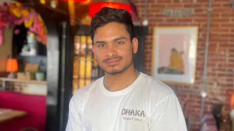 Man wearing a white t-shirt with Dhaka Restaurant branding smiling at the camera.