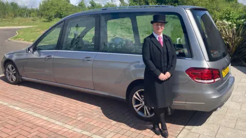 BBC/Amy Walker Rhia Carter in front of a hearse at Wealden Crematorium