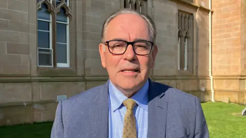 Professor Frank Casey. He has greyish receding hair, wearing black glasses a blue blazer, light blue shirt and yellow tie. 