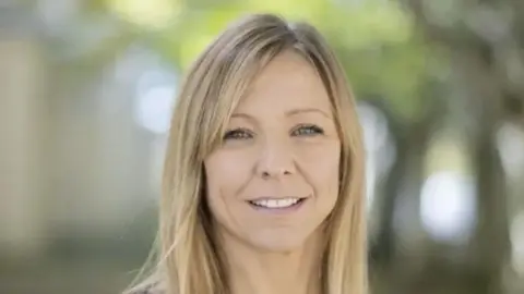 UNIVERSITY OF PLYMOUTH Close up of woman's face, she has blonde hair and is smiling while stood outside with blurred leaves in the background.