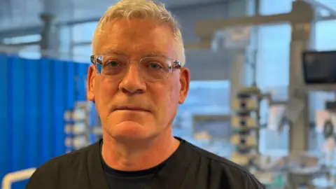 Dr Jack Parry-Jones - a man with cropped white hair and clear-rimmed glasses looks at the camera, as he stands in front of a slightly blurred hospital bed and ward in the background. 