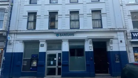 A wider look at  the exterior of the Barclays branch in Ramsey featuring a blue sign with the name of the bank. The walls are painted blue and white, and there is a brown wooden door, clear double doors, and features a cash point machine.