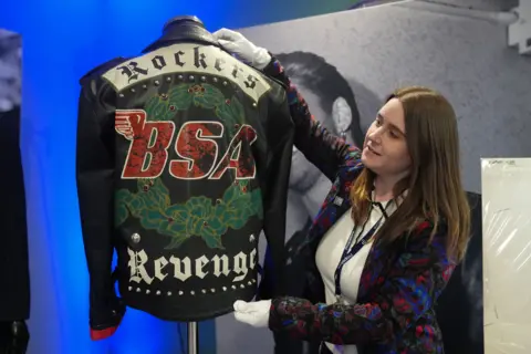 PA Media A woman is holding the George Michael jacket while wearing white protective gloves. The jacket is black with "Rockers" "BSA" and "Revenge" written on the back of it. 