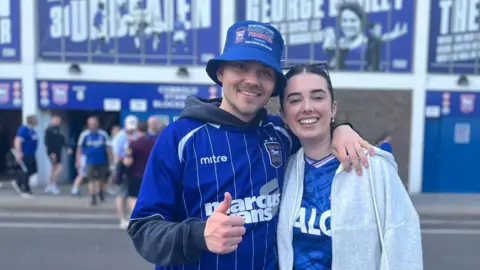 Jack Maclean/BBC Joe and Ellie and standing in front of Portman Road stadium. They are both wearing Ipswich Town FC T-shirts which are blue and white. Joe, who is on the left, is also wearing a blue bucket hat. 