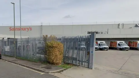 A long metal fence with a line of Sainsbury's delivery vans behind and a long building with Sainsbury's written on the side in large orange lettering