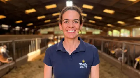 A women with black curly hair tied back, she is wearing a navy polo top with Wrights Dairy embroidered into it. She is stood in a cow shed with various cows behind her feeding. 