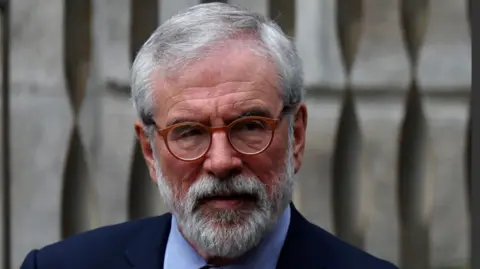 Gerry Adams has grey hair swept to the side. He is wearing red round glasses and has a beard. He is in a navy suit and blue shirt. He is looking away from the camera.