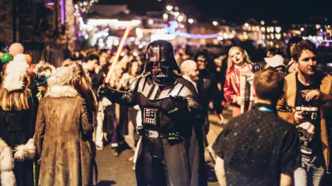 A person dressed as Darth Vader from Star Wars stands in a street full of people 