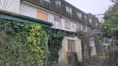 The outside of a derelict hotel in Guernsey which is boarded up and completely overgrown by greenery. 