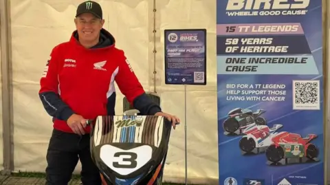 Mannin Cancers Support Group John McGuinness wears a red jumper and black cap, he stands next to a model honouring the machine he rode in 2007.