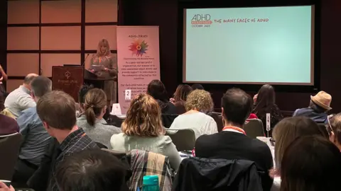 BBC A conference setting with attendees seated at round tables facing a stage. On the stage, a speaker stands near a podium with a banner that reads “ADHD Jersey” and a large screen displaying the title “The Many Faces of ADHD.” The room has modern lighting and wood-paneled walls.