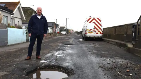 Tendring District Council A man standing next to a pothole filled with water. There is a highways maintenance van parked at the side of the road and houses on the other side. It is overcast above.