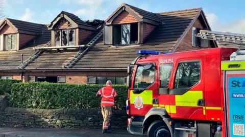 A firefighter is walking in front of the bungalow. Fire damage including broken windows can be seen at the front of the house, which is a dormer bungalow. Ladders can be seen on the roof near to the windows. A fire engine is in front of the property.