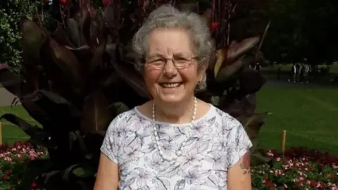 Chris Hopkins Tricia, wearing a white necklace, flowered top, with short grey hair, smiling, looking at the camera, with glasses on. She is standing in front of a large plant. 