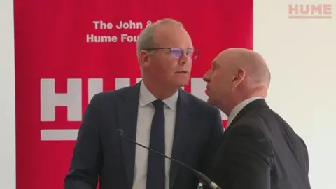 John and Pat Hume Foundation A man stands in front of a podium microphone in a navy suit and tie. He is looking off to the side as another man in a black suit leans in from the right to whisper in his ear. Behind the men is a large red sign, only partly visible, which bears the name "HUME" in capital letters. The man at the microphone has greying hair and glasses. The other man has no hair and is slightly shorter in height.