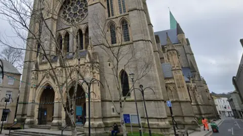 A google street view image of Truro Cathedral. It is the front of the Cathedral. The Cathedral is tall and made of a grey brownish brick. There are trees in front of it with string lights. 