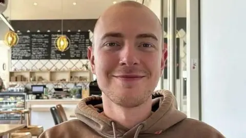 Eli smiles at the camera in a cafe. He has very short haven hair, light short stubble and dark eyebrows. He wears a brown hoodie, and has a nose ring. 
