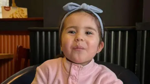 Handout Louisa "Lulu" Palmisano, three, wearing a pink fleece and a blue bow in her brown hair, sits on a black leather chair in a restaurant and smiles at the camera.