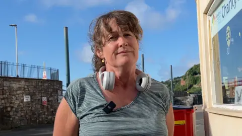 A woman wearing a grey striped top with headphones hanging around her neck. She is standing outside a train stop with blue skies behind.