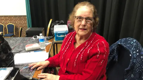 Pauline Steel is wearing a bright red sequinned cardigan.  She is wearing glasses and looking at the camera.  In front of her she has some CDs and a CD player, as she is running a dance.  She is sitting in front of a desk on the stage of a village hall where the event is taking place.  There is a black curtain behind her on the stage and boxes containing more of her CDs.