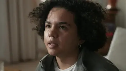 Keira Bell looking to the left. She has bobbed brown curly hair and is wearing a white t-shirt and green sweatshirt jacket