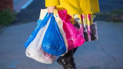 A woman carries shopping in plastic carrier bags. The woman is holding about five bags in one hand. She is wearing a floral skirt with a yellow jacket. 