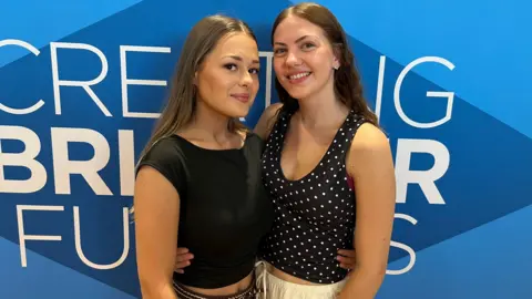 Two young women smile widely as they stand next to each other with arms wrapped around each other