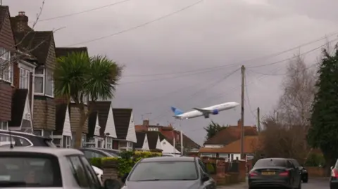 A row of suburban houses with a plane taking off very close to the houses. There are cars in the road in front of the houses. 