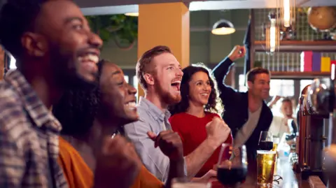 A group of people sitting a bar, cheering and punching the air. They are watching a sporting event.