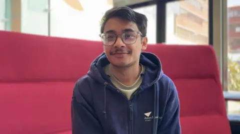 A young man in a navy hoodie, beige t-shirt and glasses sits on a red quilted sofa. He is smiling and looking slightly to the right of camera. He has short brown hair swept to the side and a slight moustache.
