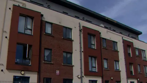 A close-up view of the exterior of a modern block of flats with brick and cream walls, tall windows and parked vehicles in the foreground