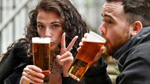 A woman and a man sitting outdoors holding pints of Lager. She is making a peace sign and he is drinking, in a casual seating area with steps and railings in the background