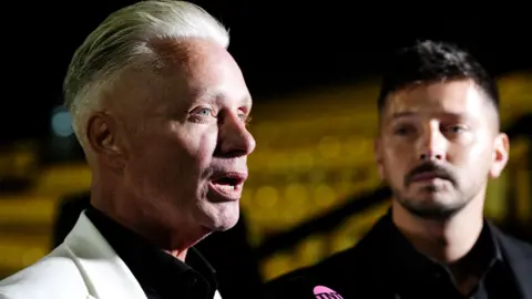 PA Media Barrie Drewitt-Barlow is being interviewed in front of a stand full of yellow seats. He is wearing a cream suit jacket over a black shirt and speaking into a microphone. Scott Drewitt-Barlow is wearing all black in the background.