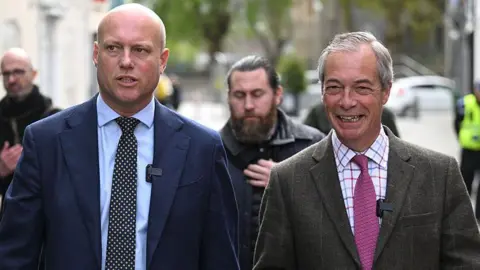 Dan Thomas is wearing a blue suit and pokadot tie with a blue shirt next to Nigel Farage, who is wearing a chequered suit, lightly chequered shirt and purple tie.