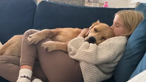 Girl Ellie lying on a sofa with golden retriever Louie lying on top of her. She has blonde hair and is wearing a beige jumper and brown leggings with  white Adidas socks. She is kissing the top of the dog's head 