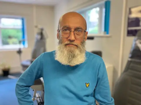 A middle-aged man with a long white beard and black glasses looks at the camera. He wears a long-sleeved blue jumper.