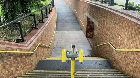 Toby Friedner/BBC A set of outdoor brick-lined steps and an adjacent sloped pathway leading down to an underpass. Yellow handrails run along both sides, with a central metal barrier dividing the stairway. The area appears damp, with scattered leaves and debris visible on the steps and at the bottom. At the far end, the entrance to the underpass features colourful graffiti on the interior wall.