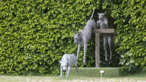 A sculpture depicts three dogs emerging from shrubs and bushes
