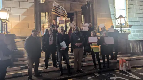 Poole Labour Thirteen men and women standing on the steps of Bournemouth Town Hall - some are holding up sheets of A4 paper but it is not possible to read what's written on them. It is after dark and the scene is lit by light shining from inside the building and large ornate lamps mounted on pillars either side of the steps